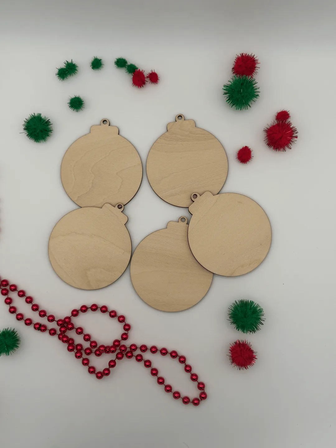 Unfinished wood christmas ornament blanks arranged in a flat lay with red garland and green and red pom poms