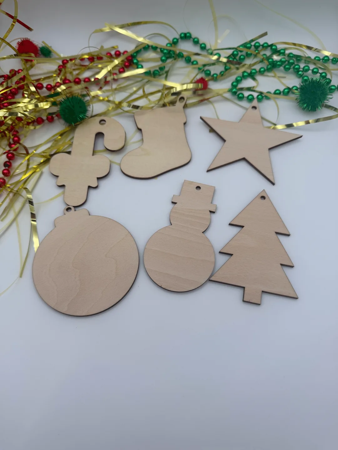 Six unfinished wood christmas ornaments in mixed holiday shapes laid flat with ribbon, pom poms, and red green bead garland