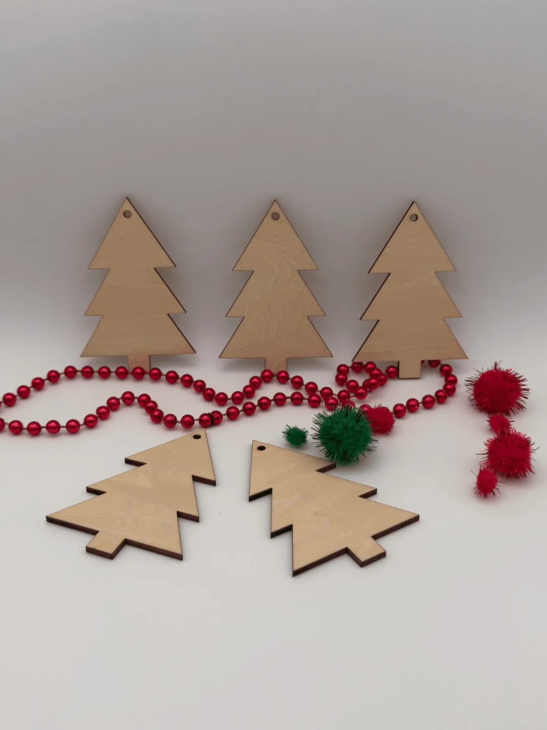Five unfinished wood christmas tree ornaments displayed upright and flat with holiday bead garland and pom poms